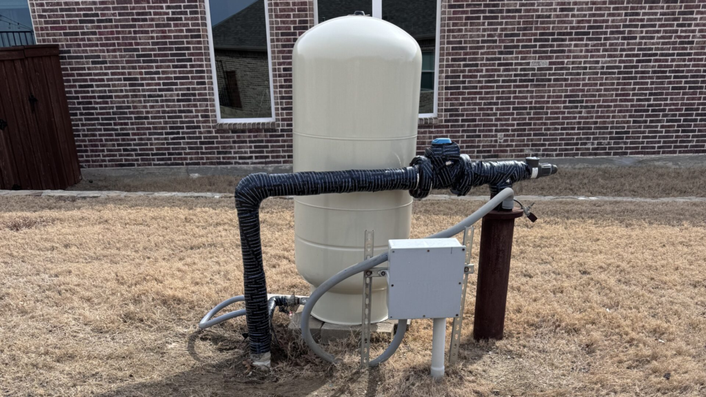 Residential water well pressure tank with insulated exterior piping beside brick home